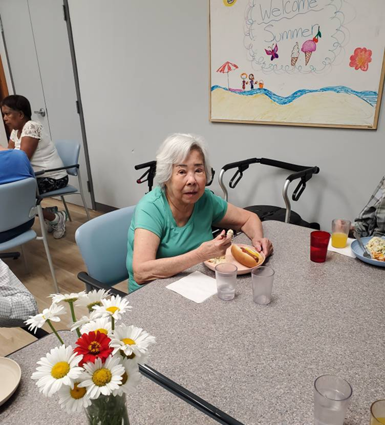 Woman eating lunch at a table with other adults in a program room with artwork on the wall.