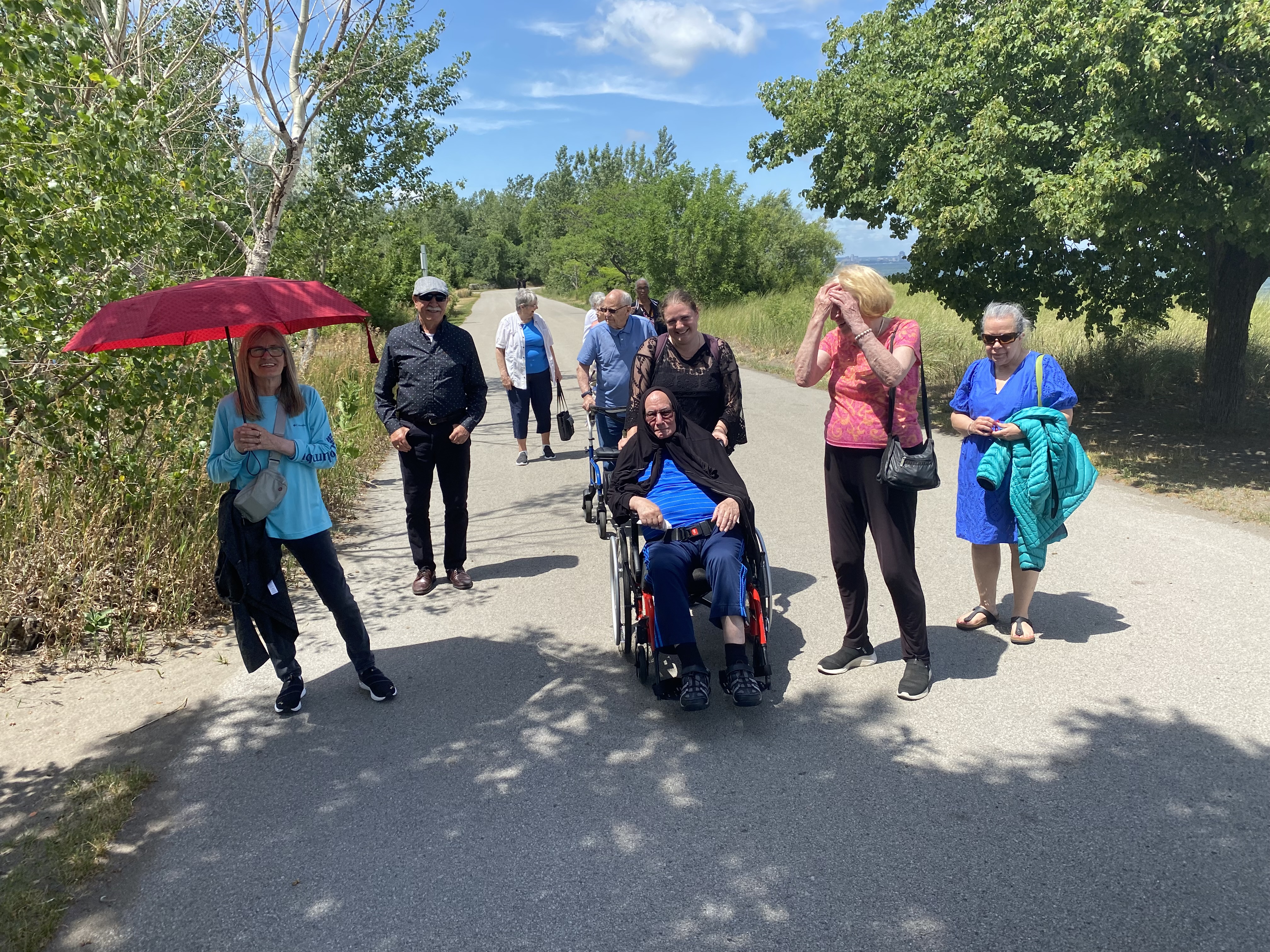 Group of adults walking together along an outdoor path, including one person using a wheelchair, during a community outing.
