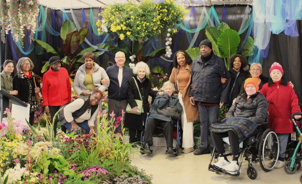 Group of adults gathered together in front of a large floral display during an indoor community outing.