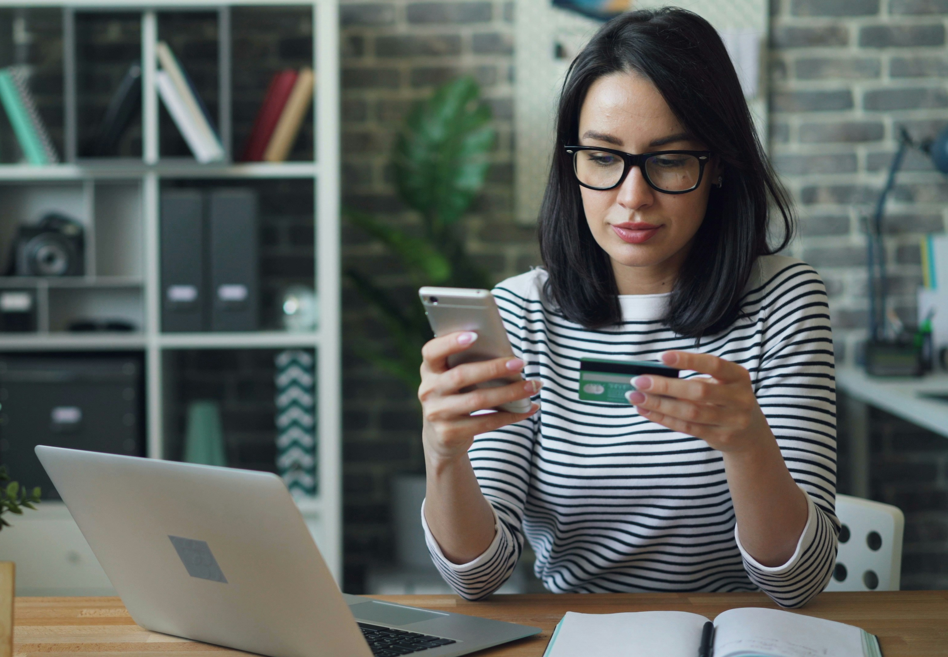 Woman sitting at a desk using her phone and holding a credit card to make an online donation.